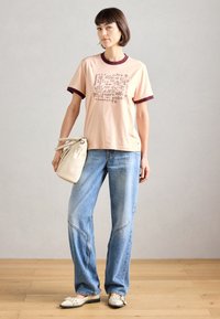 Ροδακινί βαμβακερό t-shirt με μπορντό ριγέ λεπτομέρειες και γραφικό σχέδιο. Ανοιχτό μπλε τζιν παντελόνι με ραφές. Κρέμα τσάντα και παπούτσια.