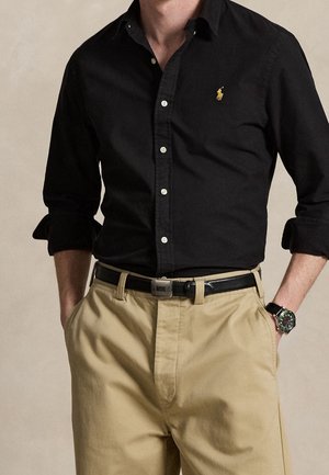 Homme portant une chemise noire boutonnée avec un logo doré, un pantalon beige avec une ceinture noire et une montre-bracelet, les mains dans les poches.