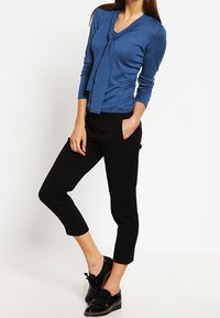 Top azul de manga larga con escote en V y detalle de nudo en el frente, combinado con pantalones cortos negros y zapatos negros con un lazo.