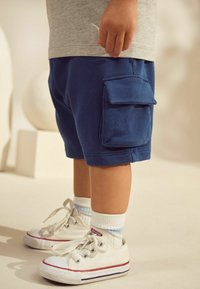 Shorts cargo en coton bleu avec une poche latérale, associés à des baskets en toile blanches et des chaussettes à rayures bleu clair. Texture lisse, design décontracté.