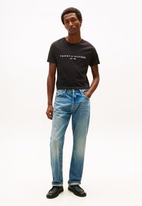 Svart bomulls-T-skjorte med "TOMMY HILFIGER"-logo i hvitt, kombinert med lyseblå denimjeans med avslappet passform og lett falming. Svarte sko.