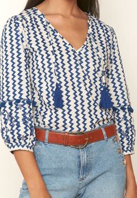 Blůza s modro-bílým cik-cak vzorem, s výstřihem do V, třásněmi a vyšívanými rukávy. Spárována s džínami s vysokým pasem a hnědým opaskem.