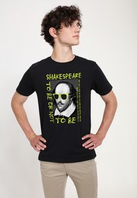 Černé bavlněné tričko s grafikou Shakespeara v žlutých slunečních brýlích, textem "SHAKESPEARE BÝT NEBO NEBÝT" a výraznými akcenty.