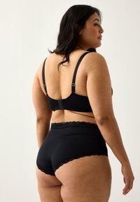 Kvinde med langt mørkt hår iført sort bh og sort undertøj med blondekant, stående med ryggen til en ensfarvet lys baggrund.