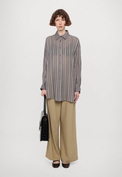Camicia sheer a righe grigie con colletto e chiusura frontale a bottoni, abbinata a pantaloni larghi beige; modella che tiene una borsa nera in tessuto testurizzato.