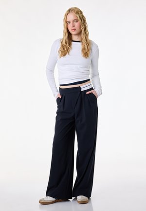 Giovane donna con lunghi capelli biondi, indossa un top corto bianco a maniche lunghe, pantaloni neri larghi e scarpe da ginnastica bianche, in piedi con le mani nelle tasche.