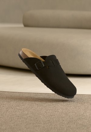 Schwarzer Slip-on-Clog mit Schnallenriemen und Korksohle, der über einem beigen Teppich vor einem beigen Sofa schwebt.