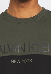 Sudadera verde y negra con cuello acanalado; presenta texto en relieve "CALVIN KLEIN" y "NEW YORK" en colores contrastantes.