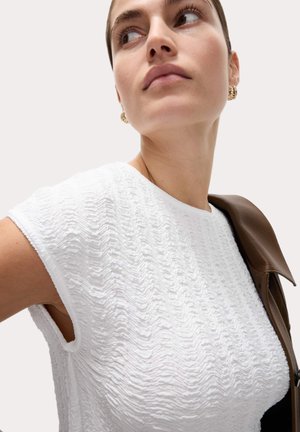 Femme portant un haut sans manches blanc texturé et des boucles d'oreilles créoles dorées, avec une veste marron posée sur son épaule, regardant vers le haut et sur le côté.