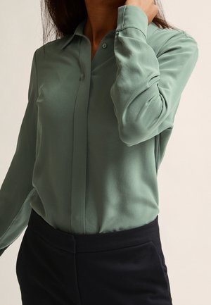 Person trägt ein langärmliges, salbeigrünes Hemd mit Knöpfen, das in hoch Taillierte schwarze Hose gesteckt ist, mit einer Hand, die den Hals berührt.