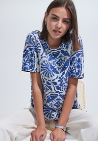 Sininen ja valkoinen kukkakuvioinen t-paita, jossa on lyhyet hihat ja pyöreä pääntie; valmistettu kevyestä kankaasta; käytetty vaaleanbeigejen housujen kanssa.