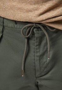 Pantalons vert olive avec une texture lisse, comprenant une fermeture à cordon de serrage avec des embouts métalliques et des poches latérales.