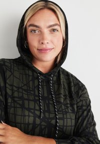 Crna in olivno zelena kapuca s potiskom v mrežo, s čimprijemniki in teksturirano blago. Ima sproščen fit s dolgimi rokavi.