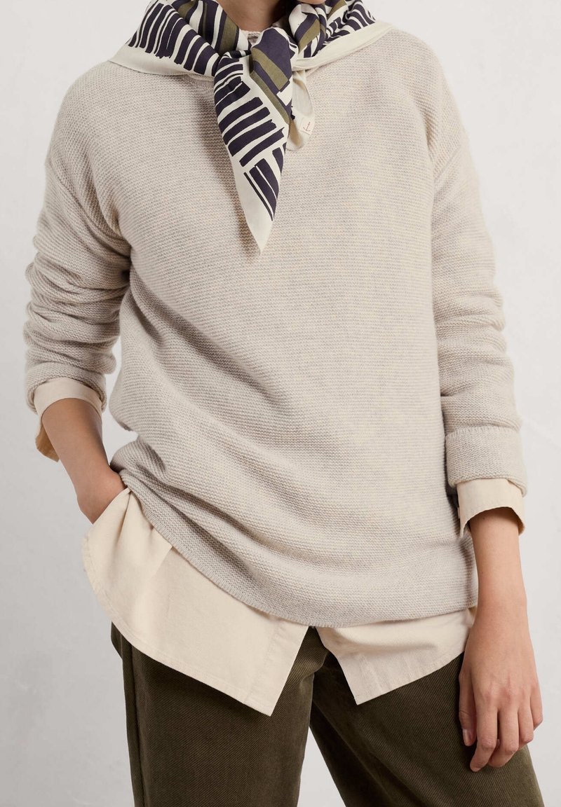 Personne portant un pull en maille beige par-dessus une chemise crème, un pantalon olive, et une écharpe rayée nouée autour du cou, main dans la poche.