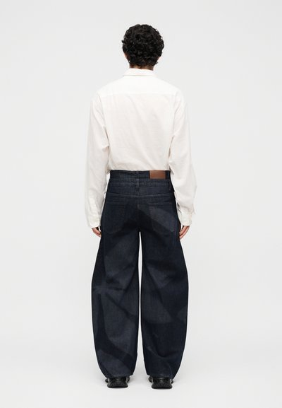 Άτομο με σγουρά μαλλιά που φοράει άσπρο πουκάμισο μακρυμάνικο και φαρδιά σκουρομπλε τζιν, στέκεται γυρισμένο από πίσω σε μονόχρωμο φόντο.