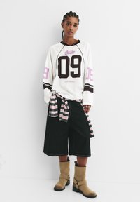 Weißes Langarmhemd mit schwarzen und rosa Akzenten, versehen mit der Nummer 09. Kombiniert mit schwarzen weiten Shorts und beige Stiefeletten. Gestreiftes Hemd um die Taille gebunden.