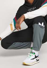 Svart hoodie med vita och färgglada randiga ärmar, matchad med svarta och grå träningsbyxor med ett Adidas-logotyp. Vita sneakers med gröna accenter.