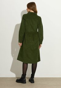 Groene corduroy jurk met een kraaglijn, lange mouwen, gathered taille en midi-lengte, gecombineerd met zwarte enkellaarsjes.