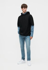 Νέος άνδρας που φοράει μαύρο κοντό μπουφάν με κουκούλα πάνω από μπλε μακρυμάνικο πουκάμισο, ανοιχτό μπλε τζιν και λευκά αθλητικά παπούτσια, στέκεται μπροστά σε λευκό φόντο.