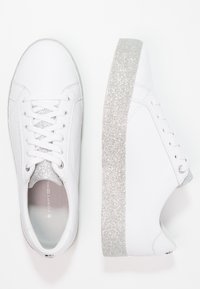 Sneakers bianchi in pelle con suola glitterata argento e dettagli in lacci, caratterizzati da una superficie liscia e design a punta rotonda.