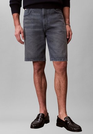 Hombre que lleva pantalones cortos de mezclilla grises, mocasines negros y una camisa negra de manga larga, de pie frente a un fondo gris liso.