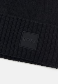 Fekete kötött sapka bordázott mintával és egy texturált, négyzet alakú gumipatchel, amelyen a "BOSS" márkaneve középen mélynyomva található.
