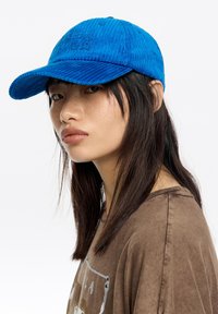 Mujer joven con cabello oscuro y liso, lleva una gorra de pana azul brillante y una camiseta marrón con diseño gráfico, mirando ligeramente de lado sobre un fondo blanco.