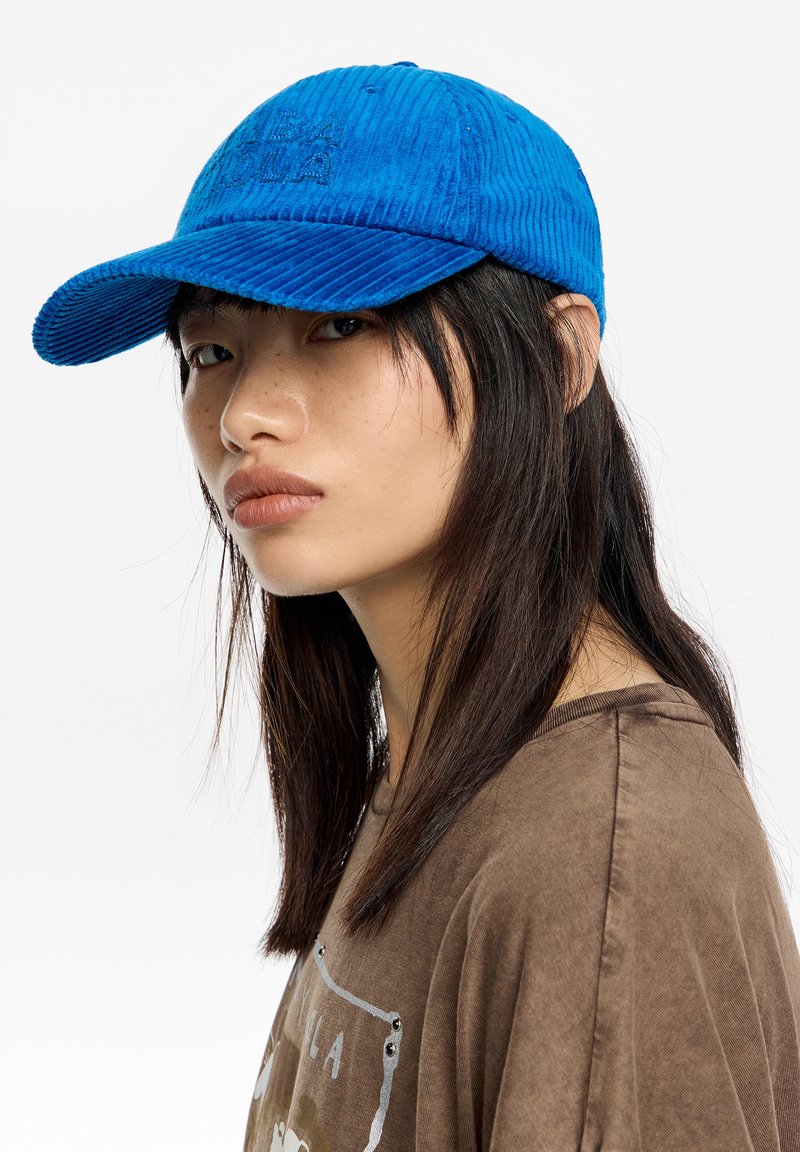 Mujer joven con cabello oscuro y liso, lleva una gorra de pana azul brillante y una camiseta marrón con diseño gráfico, mirando ligeramente de lado sobre un fondo blanco.