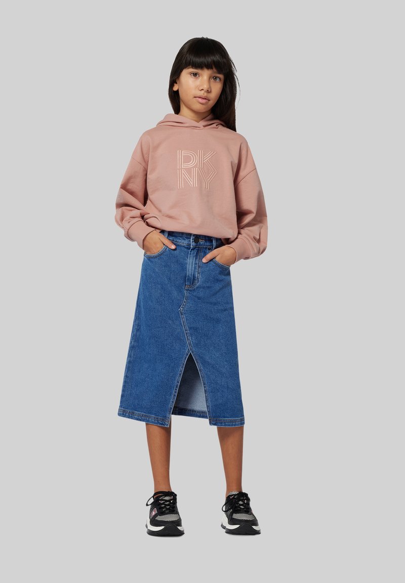 Sweatshirt à capuche rose avec logo blanc, associé à une jupe en denim bleue jusqu'aux genoux avec une fente avant et portée avec des chaussures de sport noires.