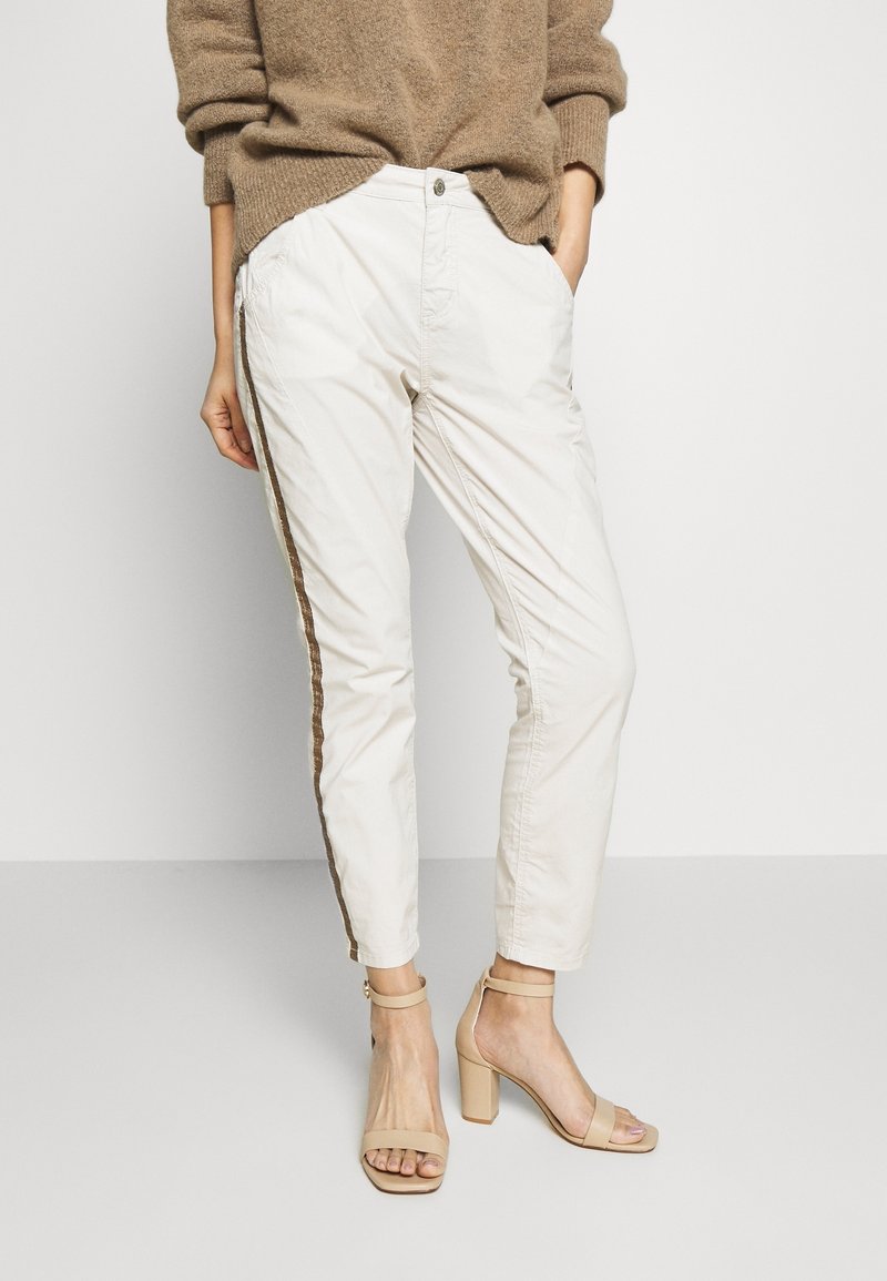 Femme portant un pull beige, un pantalon blanc longueur cheville avec une bande latérale marron, et des sandales à talons blocs couleur nude, debout les mains dans les poches.