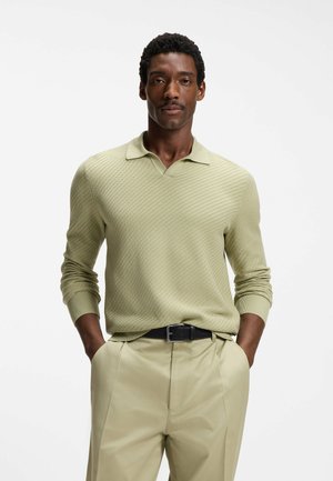 Mann mit kurzen lockigen Haaren, der ein strukturiertes hellgrünes Langarm-Poloshirt und beige Hose mit schwarzem Gürtel trägt, Hände in den Taschen.