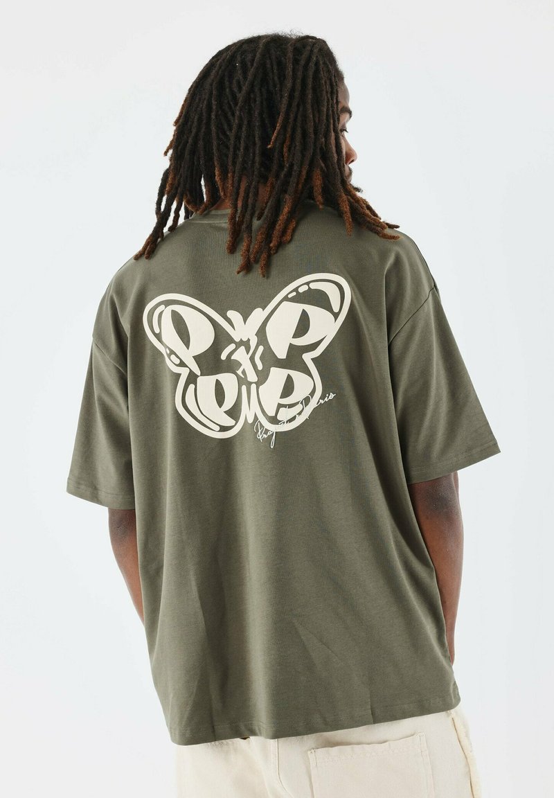 Camiseta oversized en verde oliva con un gran gráfico de mariposa blanca en la parte posterior, que presenta detalles curvados y un acento escrito a mano.