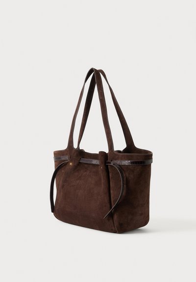 Sac cabas en daim marron foncé avec deux longues anses et de fines lanières décoratives sur un fond clair uni.