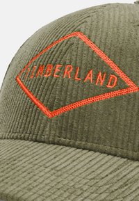 Gorra de pana verde con un logo de diamante naranja. Presenta una superficie texturizada y el nombre de la marca "TIMBERLAND" bordado en letras negritas.