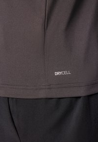 Camisa deportiva de manga corta en gris oscuro con tejido texturizado. Presenta el logo blanco "DRYCELL" cerca del dobladillo. Pantalones cortos negros parcialmente visibles.