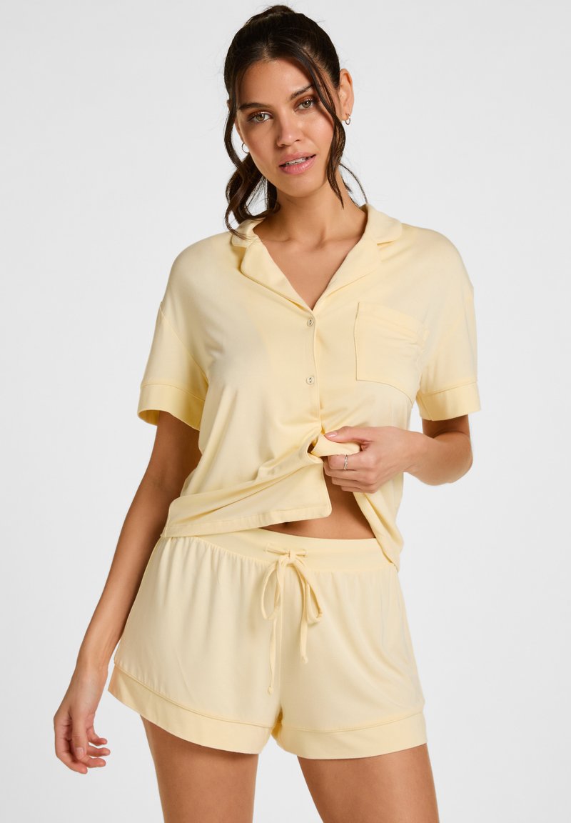 Vrouw draagt lichtgele korte mouwen blouse met knopen en bijpassende korte broek met trekkoord in de taille, poseert tegen een effen achtergrond.
