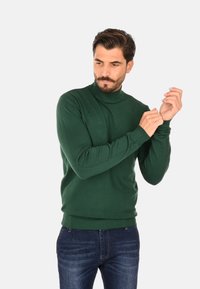 Maglione verde a collo alto in maglia con polsini e orlo a coste, abbinato a jeans blu. Il tessuto appare liscio, con una consistenza uniforme e senza motivi visibili.