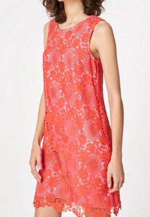 Rød ærmeløs blondekjole med blomsterprint båret af en person, der står foran en ensfarvet hvid baggrund.