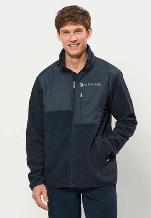 Homme souriant portant une veste zippée bleu marine U.S. Polo Assn. les mains dans les poches, sur un fond clair uni.