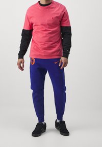 Rosa Kurzarm-T-Shirt über einem schwarzen Langarm-Oberteil, kombiniert mit blauen Jogginghosen mit orangefarbenem Nike-Logo und niederländischem Löwenemblem. Schwarze Sneakers.