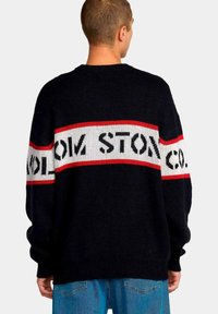 Suéter de punto azul marino con una banda horizontal roja y blanca que presenta un texto en negrita. Corte ligeramente oversized con mangas largas y cuello redondo.
