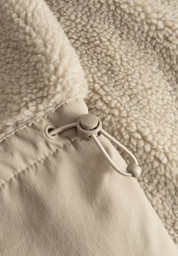 Beige fleece stof med en tekstureret overflade, der har en glat, lys beige foring og et justerbart snoretræk i kanten.