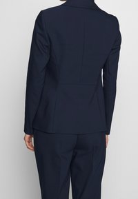 Námořnický blazer s přiléhavým střihem, který má klopový límec, detaily na zadním švu, hladkou texturu látky a strukturovanou siluetu.