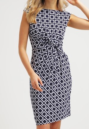Frau trägt ein ärmelloses marinefarbenes Kleid mit weißen geometrischen Quadratmustern und einem gerafften Knotendetail an der Taille.