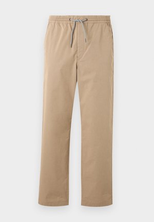 Ung mand stående med hænderne i lommerne iført en sort t-shirt med fire farverige kraniedesigns, beige bukser og hvide sneakers.