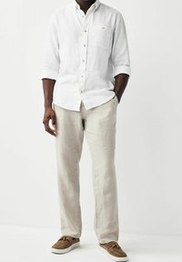 Uomo che indossa una camicia bianca a maniche lunghe con bottoni, pantaloni beige chiaro e mocassini marroni con nappe, in piedi con una mano in tasca.