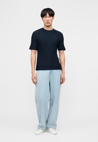 Άνδρας που φοράει υφασμένο μαύρο πουλόβερ με κοντά μανίκια, ανοιχτό μπλε τζιν παντελόνι φαρδύ, και λευκά αθλητικά παπούτσια, στέκεται μπροστά σε ένα απλό ανοιχτό φόντο.