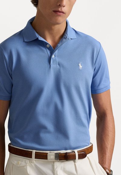 Un tricou polo de culoare albastru deschis, cu un material texturat, cu o deschidere cu două butoane și un mic logo alb pe piept. Purtați-o cu o curea din piele maro.