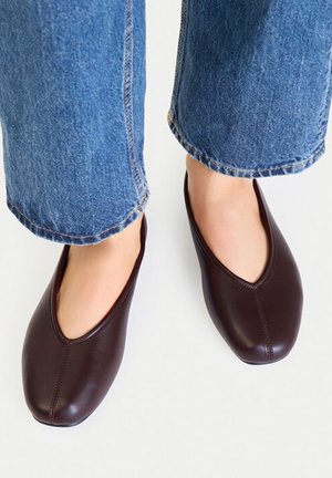 Persona que lleva zapatos de cuero marrón oscuro sin cordones con punteras redondeadas y vaqueros azules contra un fondo claro y liso.