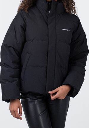 Chaqueta acolchada negra Carhartt con cuello alto, bolsillos laterales, llevada por una persona con pantalones de cuero negro, mano ajustando el frente de la chaqueta.